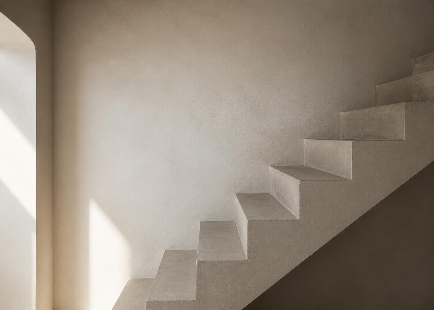 A bare concrete staircase rising against a pale plaster wall in soft daylight